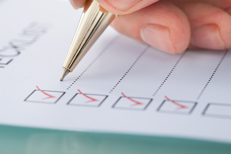 A hand holds a gold pen, marking off items on a checklist printed on white paper, indicating preparation tasks related to post weight-loss surgery.