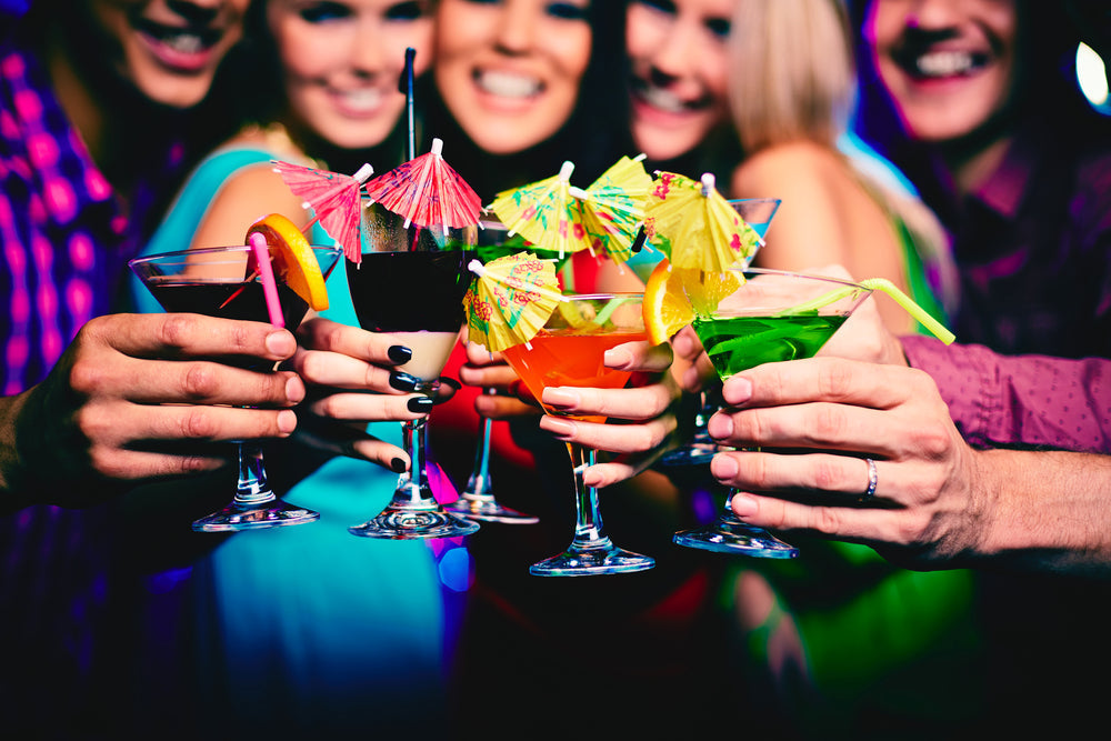 A group of five friends joyfully clinks colorful cocktails and mocktails adorned with umbrellas in a vibrant setting, celebrating together.