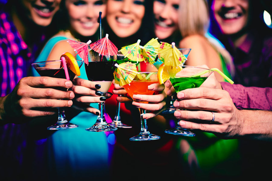 A group of five friends joyfully clinks colorful cocktails and mocktails adorned with umbrellas in a vibrant setting, celebrating together.