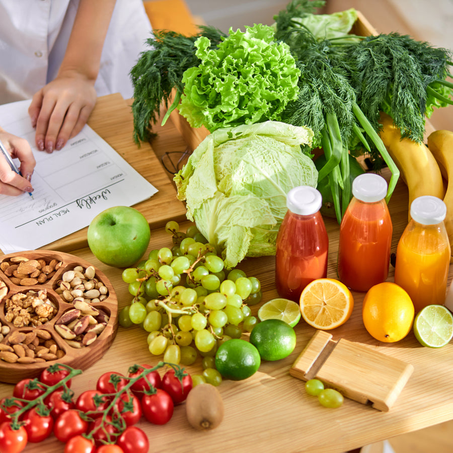 A person writes on a meal plan sheet while surrounded by fresh fruits, vegetables, nuts, and colorful juices on a wooden table, emphasizing healthy eating for bariatric diet stages.