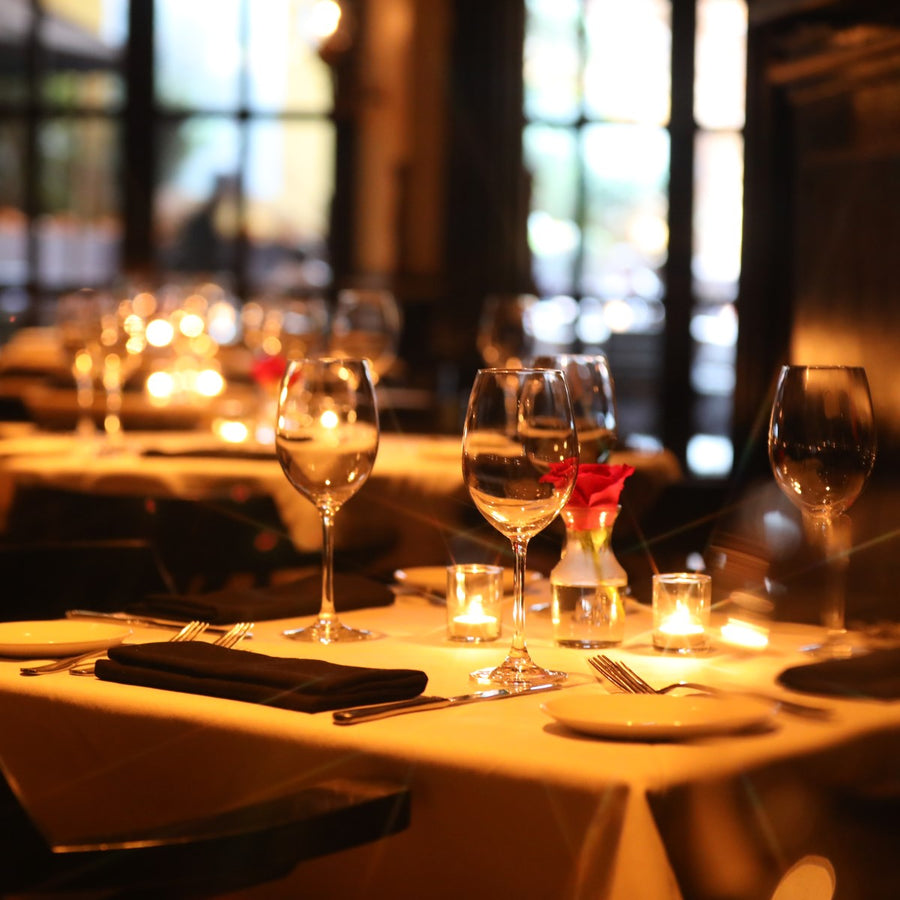 A dimly lit restaurant table is elegantly set with wine glasses, silverware, and a small vase of roses, creating a warm and inviting atmosphere for dining.