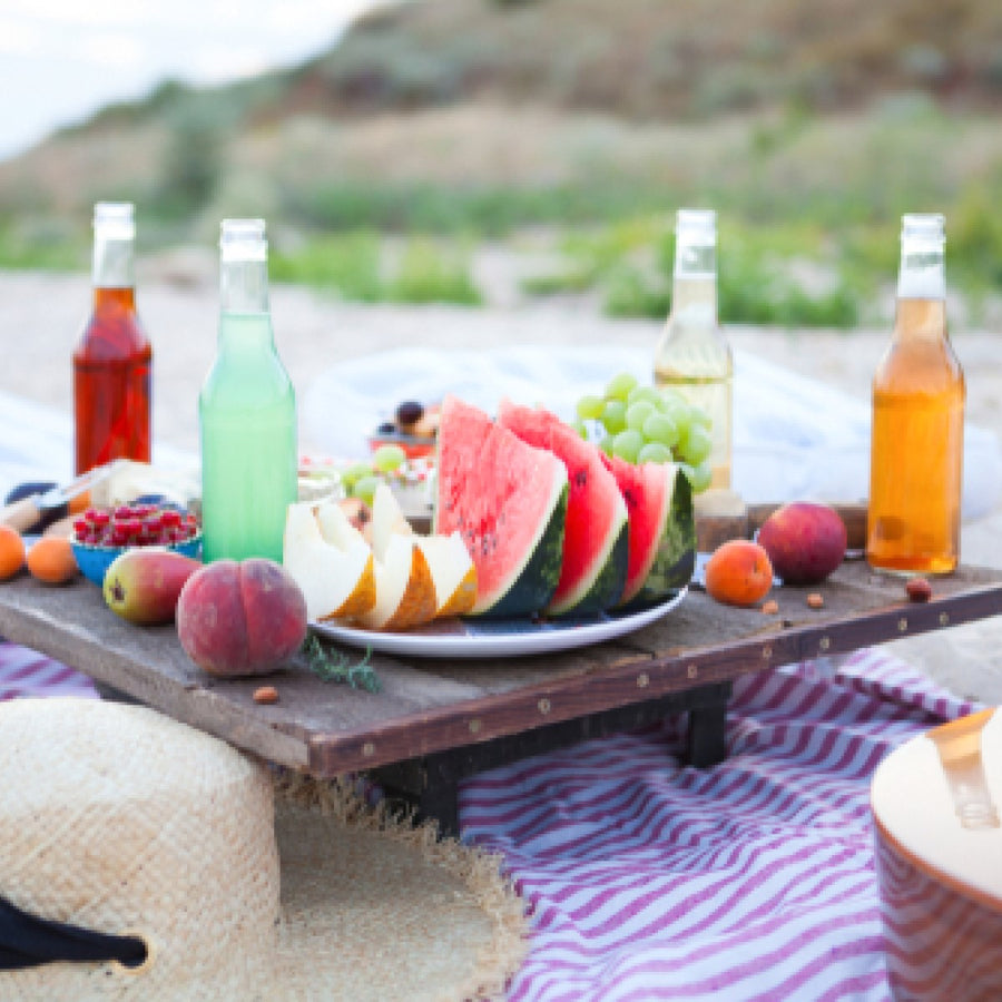A picnic table displays a variety of fresh fruits, including watermelon, grapes, and peaches, alongside colorful drinks, set against a natural outdoor backdrop.