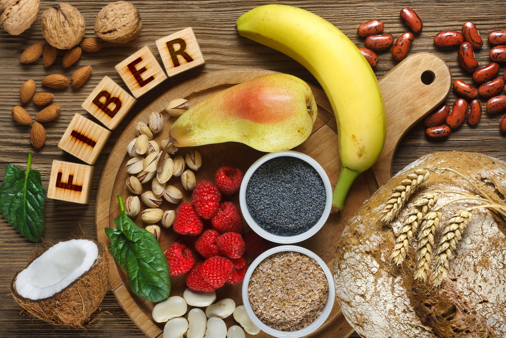 A wooden platter displays various fiber-rich foods, including a banana, pear, nuts, seeds, raspberries, and whole grain bread, with the word "FIBER" spelled out in letter blocks.