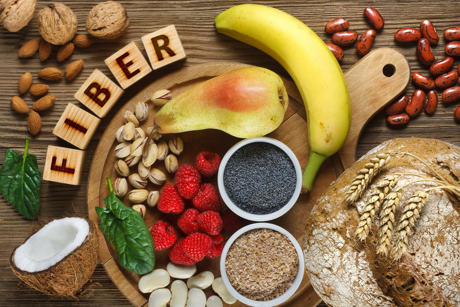 A wooden platter displays various fiber-rich foods, including a banana, pear, nuts, seeds, raspberries, and whole grain bread, with the word "FIBER" spelled out in letter blocks.