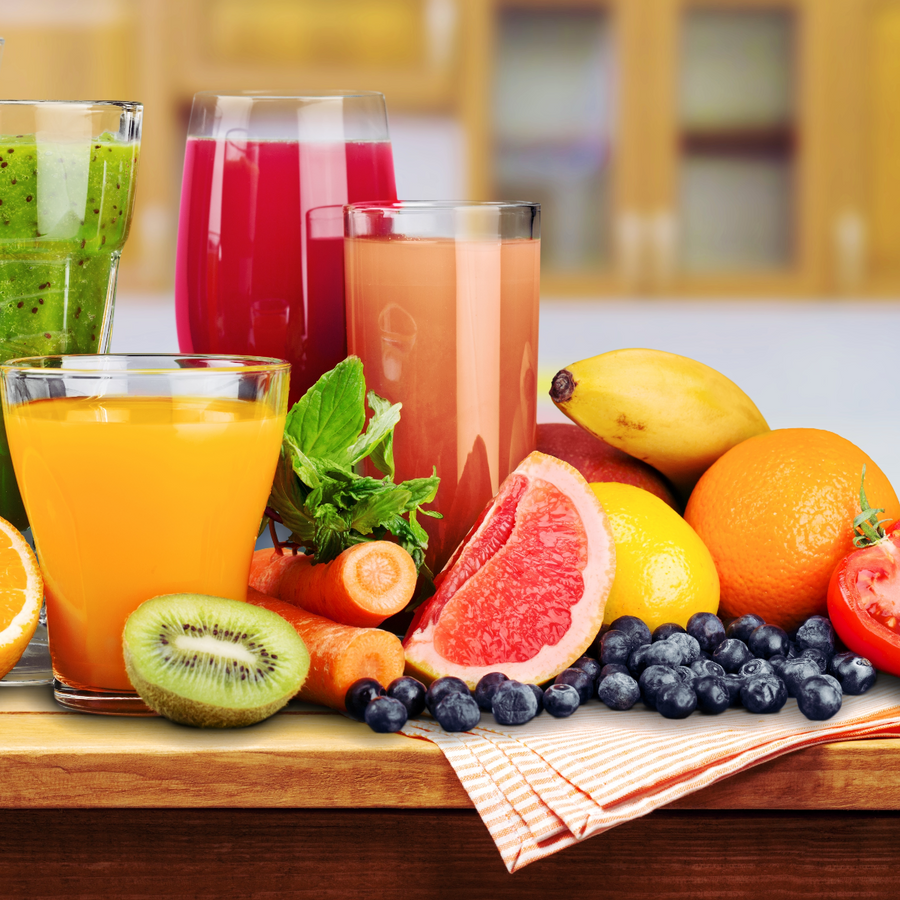 A variety of colorful drinks, including orange juice, a green smoothie, and fruit juices, are displayed alongside fresh fruits and vegetables like bananas, oranges, and carrots on a wooden table in a kitchen setting.