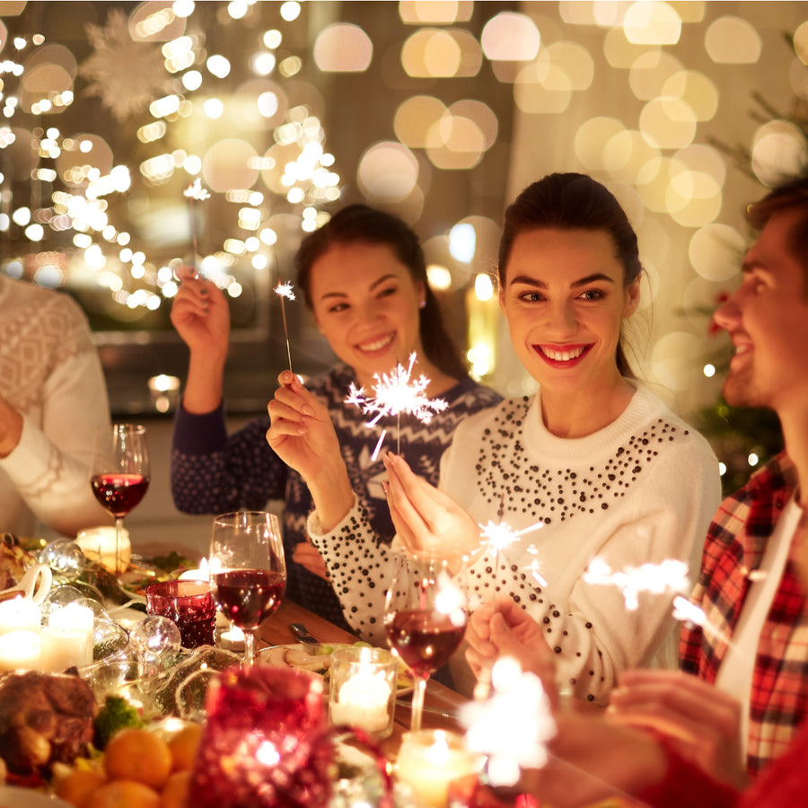 A group of four smiling friends at a festive holiday table hold sparklers, surrounded by candles and decorations, enjoying a joyful gathering with food and drinks in a warmly lit setting.