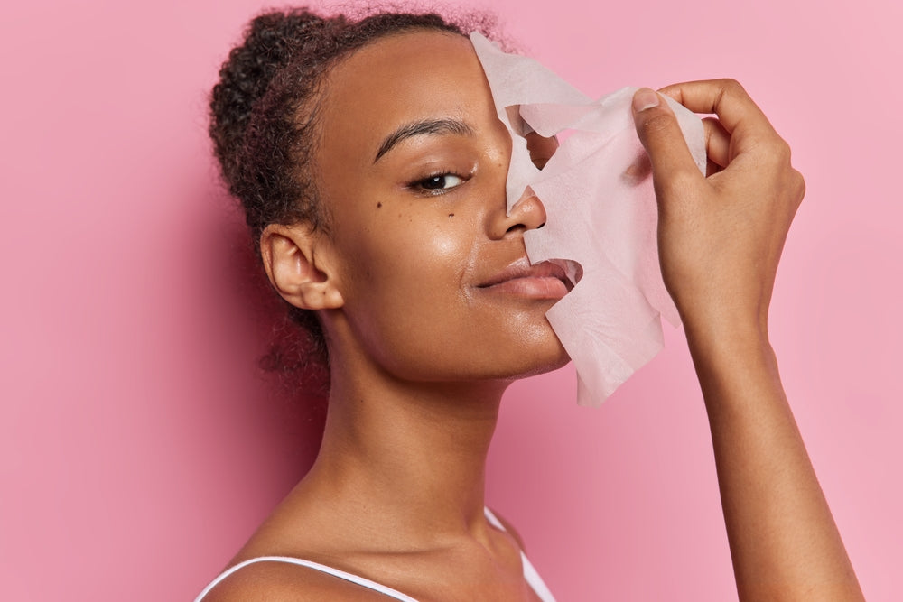 A woman with curly hair smiles while holding a facial mask away from her face, against a bright pink background, showcasing a skincare routine.