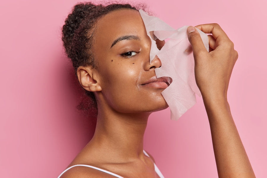 A woman with curly hair smiles while holding a facial mask away from her face, against a bright pink background, showcasing a skincare routine.