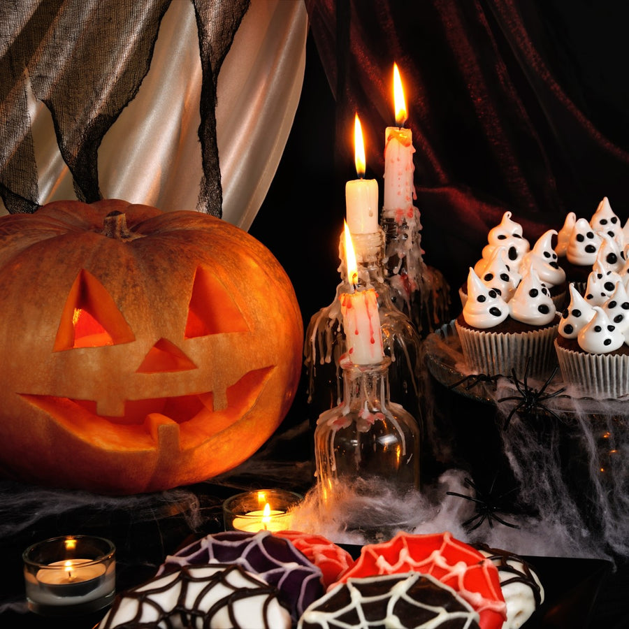 A carved jack-o'-lantern glows beside spooky cupcakes topped with ghostly frosting, while lit candles and Halloween-themed cookies create a festive, eerie atmosphere.