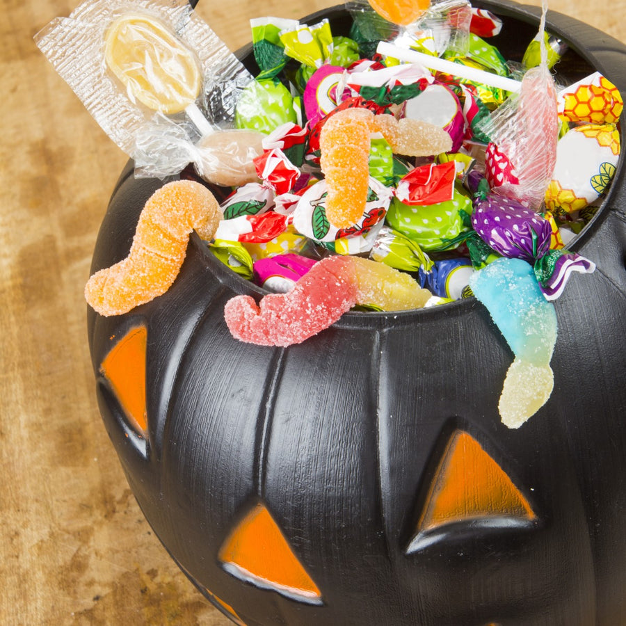 A black pumpkin-shaped bowl filled with colorful Halloween candies, including gummy worms and lollipops, sits on a wooden surface, showcasing bariatric-friendly treats for the holiday.