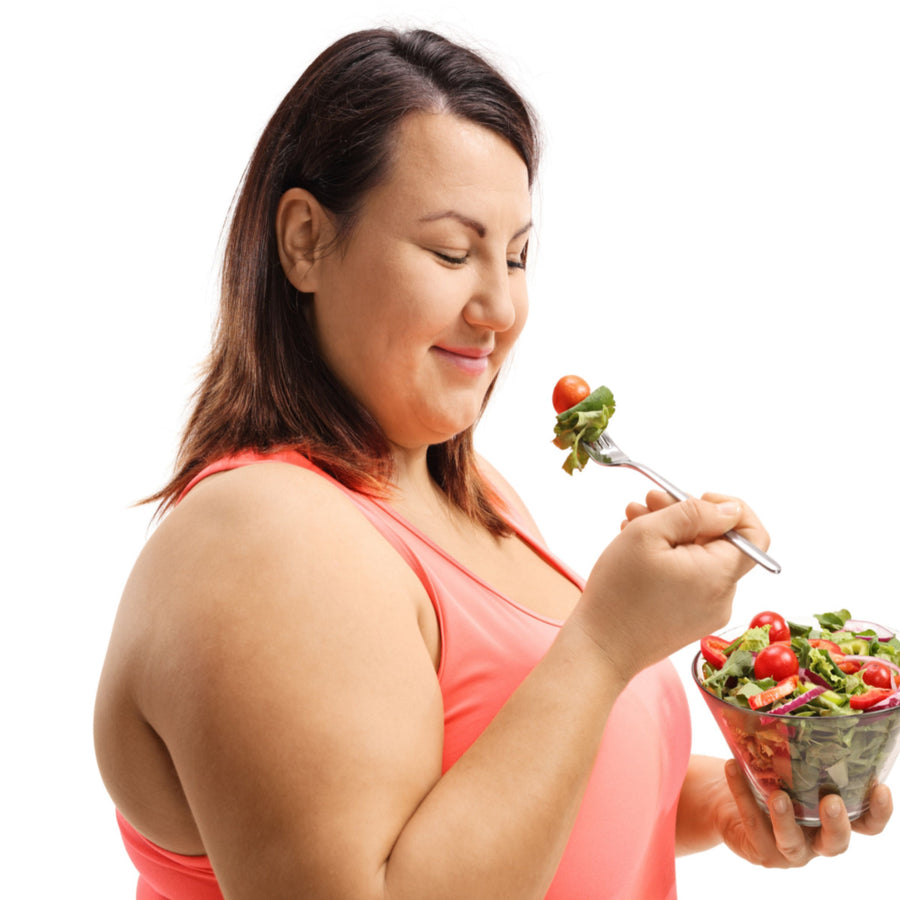 A woman in a pink tank top smiles as she holds a fork with a salad, looking content while preparing to eat from a bowl of fresh greens and cherry tomatoes.