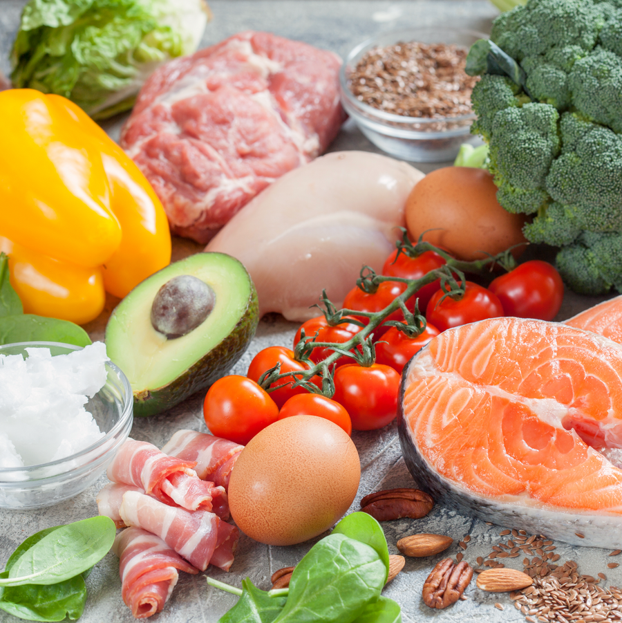 A variety of fresh ingredients including yellow bell pepper, meats, avocado, cherry tomatoes, broccoli, eggs, and nuts are arranged on a gray surface, emphasizing healthy options for post-bariatric surgery meals.