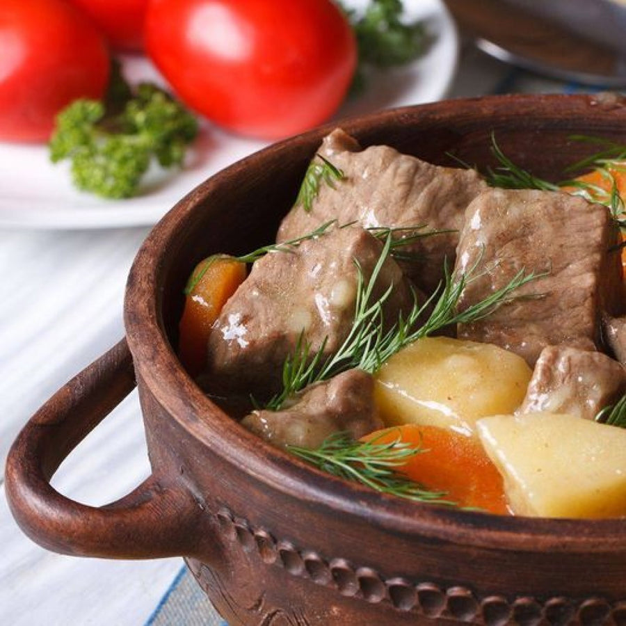 A wooden bowl contains a hearty stew with chunks of beef, potatoes, and carrots, garnished with fresh dill, alongside ripe tomatoes and parsley in the background.