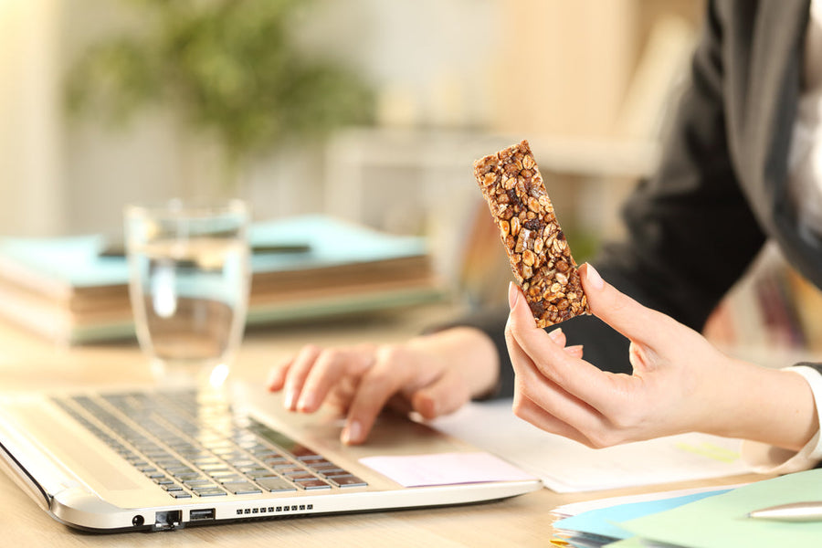 A person in business attire holds a granola bar in one hand while typing on a laptop with the other, surrounded by a glass of water and colorful files in a bright office setting.
