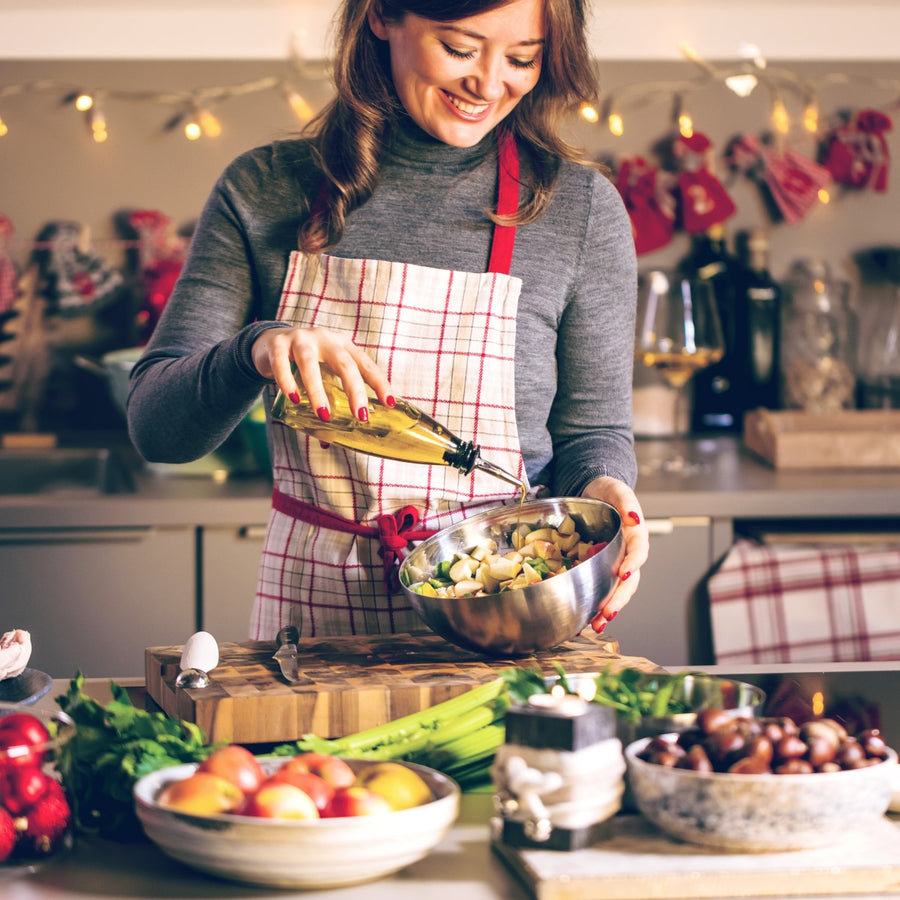 A woman in a gray sweater and plaid apron smiles as she drizzles olive oil into a bowl of chopped vegetables while preparing food in a festive kitchen decorated with lights.
