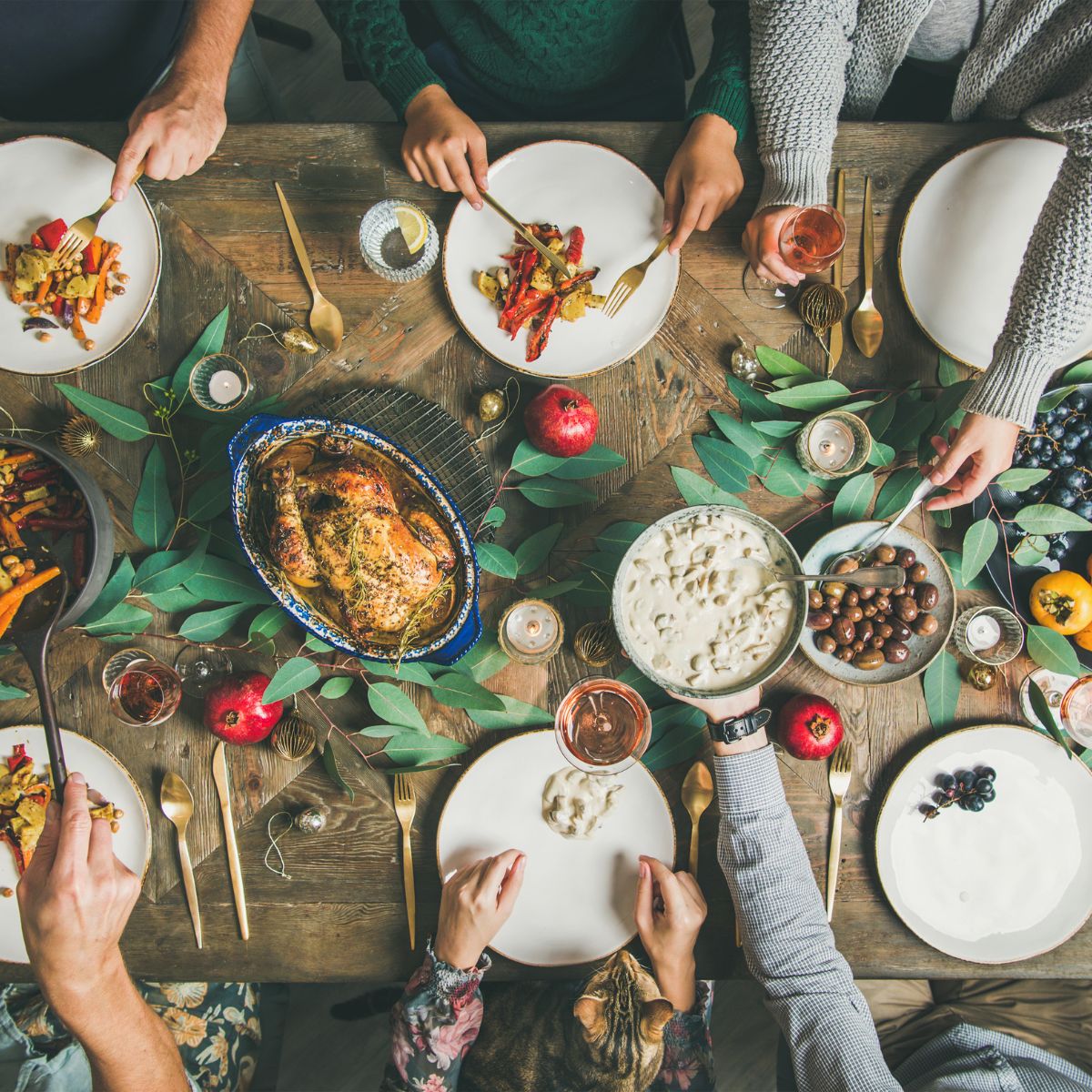 A festive table is set with plates of food, including a roasted chicken, vegetables, and desserts, as people enjoy a meal together, surrounded by greenery and candles, with a cat nearby.