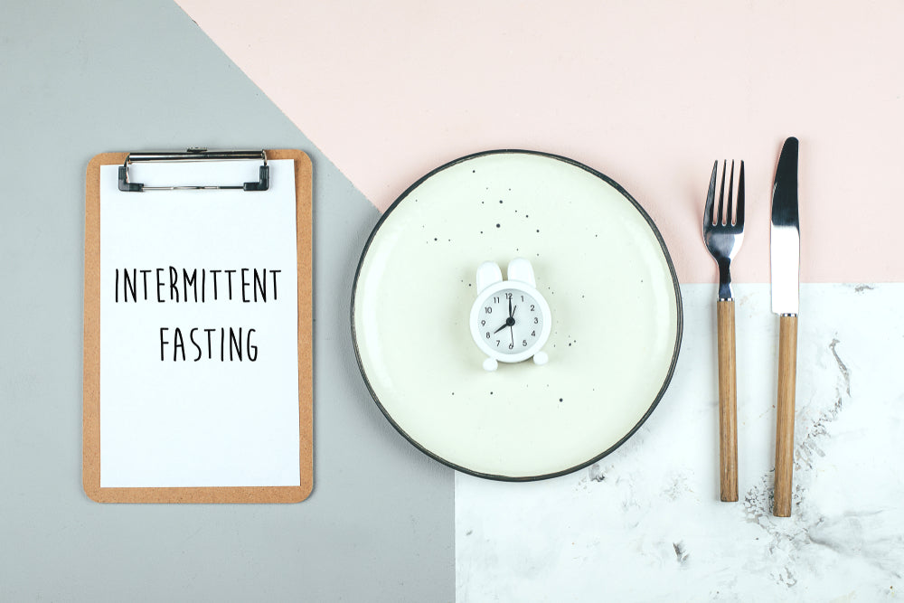 A clipboard with "Intermittent Fasting" written on it sits next to a plain plate with a small white alarm clock and a knife and fork, set against a pastel-colored background.