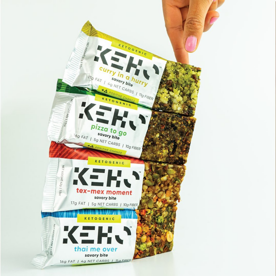 A hand holds a stack of four KEHO Savory Keto Bites packages labeled "curry in a hurry," "pizza to go," "tex-mex moment," and "thai me over," showcasing their ketogenic snack options against a white background.