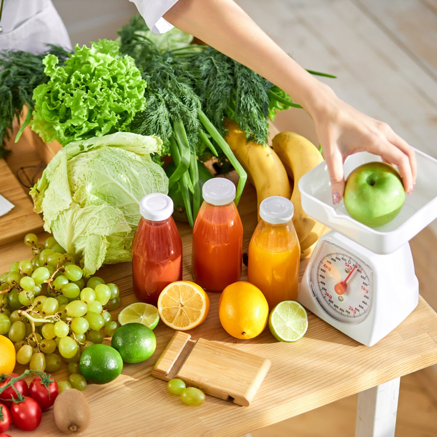 A hand places a green apple on a kitchen scale surrounded by fresh fruits, vegetables, and colorful juice bottles on a wooden table, emphasizing healthy eating after bariatric surgery.