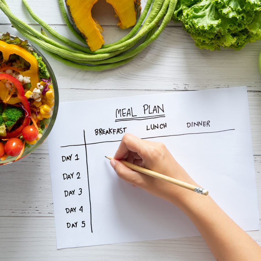 A hand holds a pencil to write on a blank meal plan for five days, with a colorful salad bowl and fresh vegetables like lettuce and squash visible in the background.