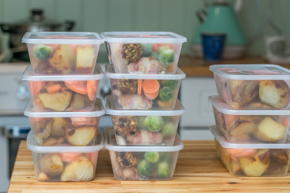 Stacked clear containers filled with colorful cooked vegetables, including carrots, broccoli, and potatoes, are arranged neatly on a wooden countertop in a kitchen setting.