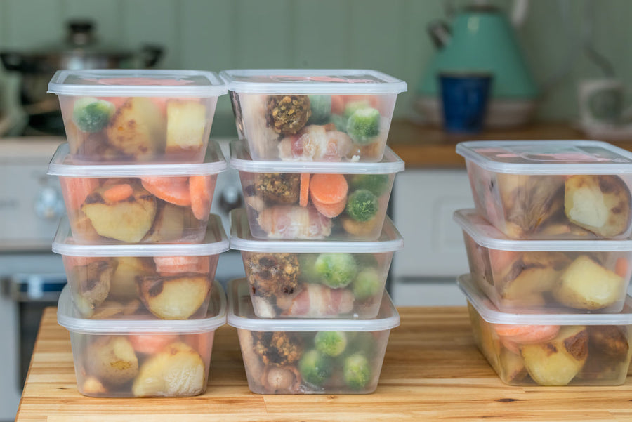 Stacked clear containers filled with colorful cooked vegetables, including carrots, broccoli, and potatoes, are arranged neatly on a wooden countertop in a kitchen setting.