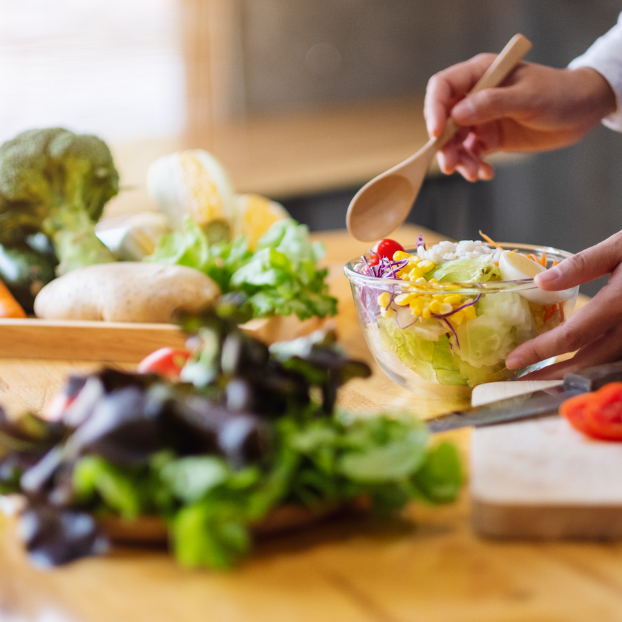 A person stirs a colorful salad in a clear bowl with a wooden spoon, surrounded by fresh vegetables on a wooden countertop, emphasizing healthy meal prep.