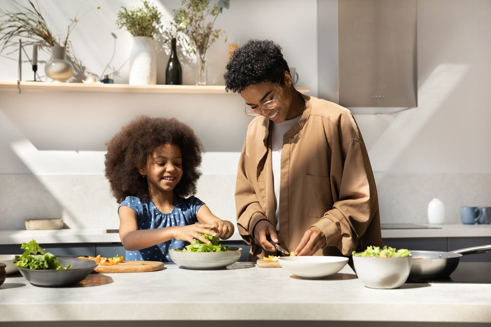 A smiling child and an adult prepare fresh vegetables together at a modern kitchen counter, surrounded by bowls of ingredients and natural light streaming in from a window.