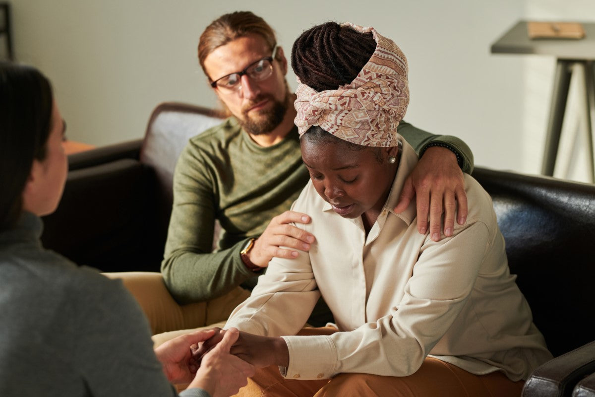 A woman with a headscarf looks down, appearing distressed while holding hands with another person, as a man gently comforts her from behind in a cozy indoor setting.