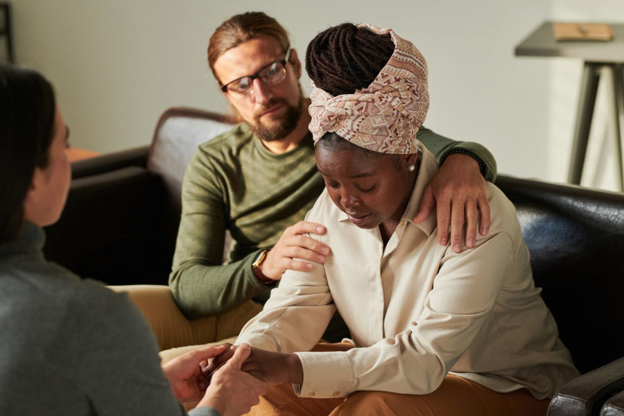 A woman with a headscarf looks down, appearing distressed while holding hands with another person, as a man gently comforts her from behind in a cozy indoor setting.