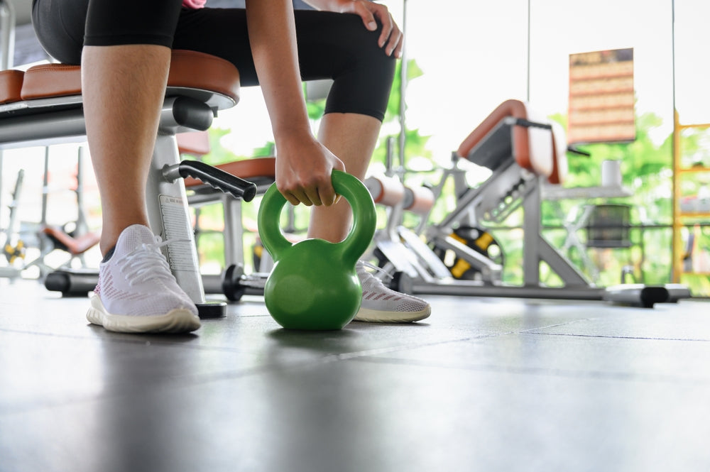 A person in athletic wear is preparing to lift a green kettlebell while seated on a bench in a bright gym filled with exercise equipment.