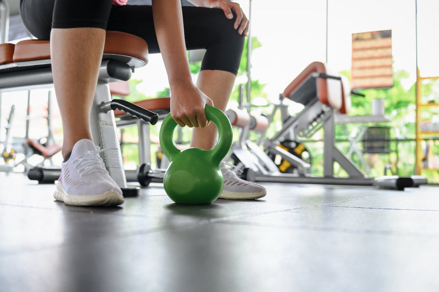 A person in athletic wear is preparing to lift a green kettlebell while seated on a bench in a bright gym filled with exercise equipment.