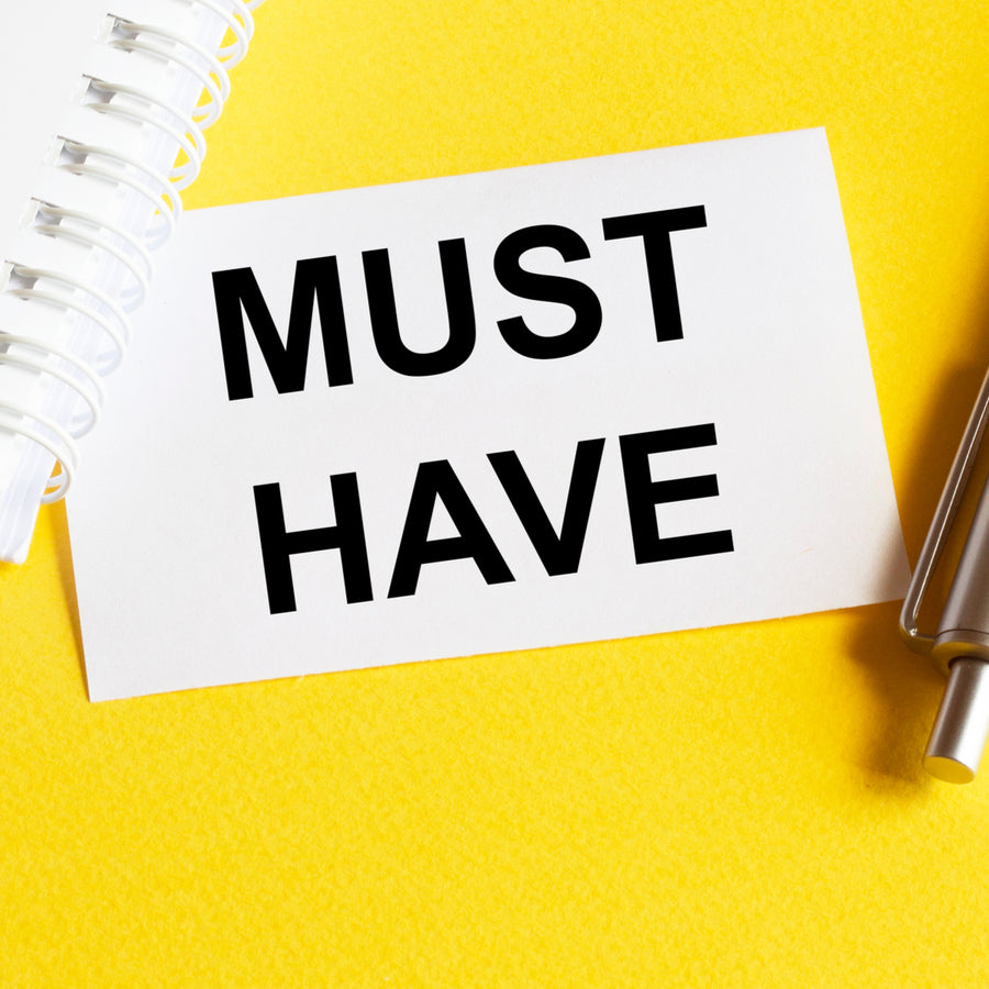 A white card with bold black text reading "MUST HAVE" is placed on a bright yellow background, alongside a spiral notebook and a silver pen, suggesting important items related to bariatric surgery.