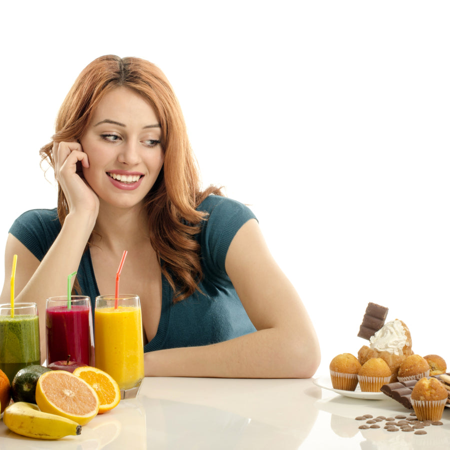 A smiling woman rests her chin on her hand, looking at a colorful array of smoothies and a plate of desserts, including cupcakes and chocolate, on a bright, white table.