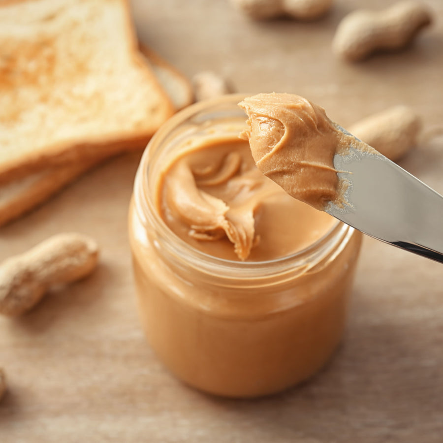 A knife holds a scoop of creamy peanut butter above a jar, with a slice of toasted bread and scattered peanuts in the background on a wooden surface.