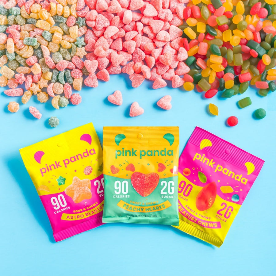 Colorful gummy candies in various shapes and sizes are scattered on a light blue background, with three brightly packaged Pink Panda candy bags labeled "Astro Blasters," "Peachy Hearts," and "Pleasure Chews."