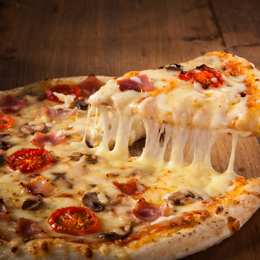 A slice of cheesy pizza topped with tomatoes, mushrooms, and ham is being lifted from the whole pizza, showcasing its gooey melted cheese against a wooden table background.