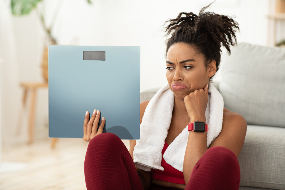A woman with curly hair sits on the floor in a living room, wearing workout clothes and a towel around her neck, looking frustrated while holding a scale.