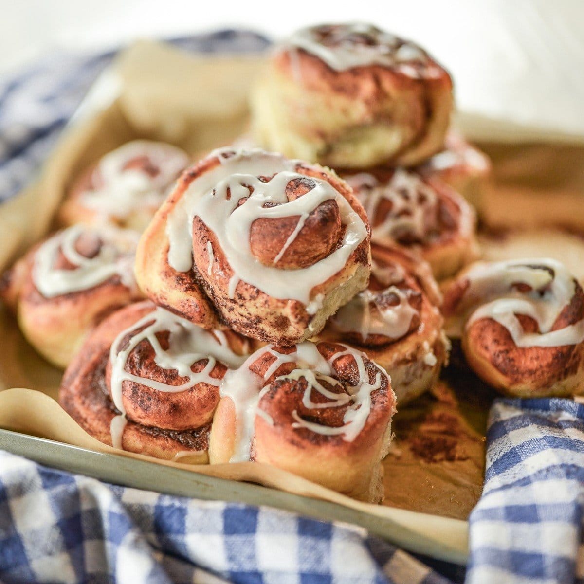 A stack of freshly baked protein cinnamon rolls drizzled with white icing sits on parchment paper, surrounded by a blue and white checkered cloth.