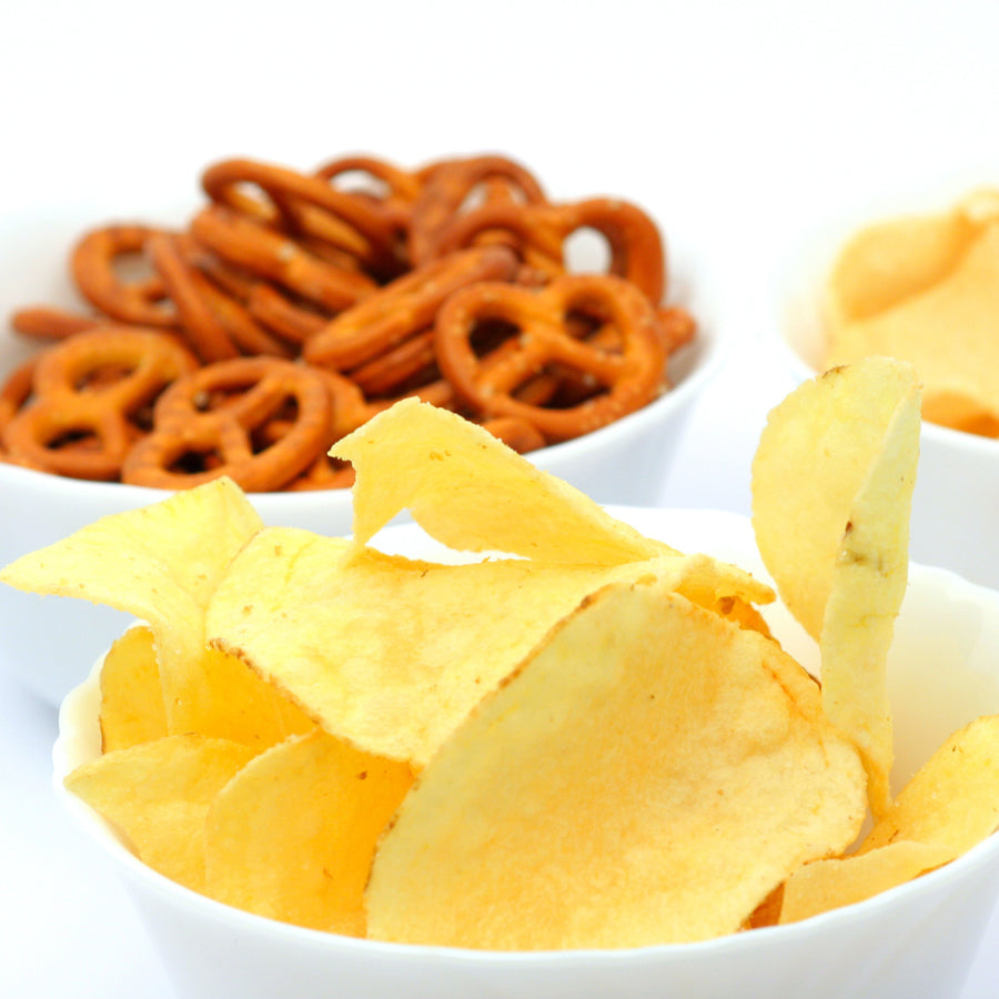 A close-up of a bowl filled with yellow potato chips, with another bowl of brown pretzels in the background, showcasing a variety of high-protein snacks suitable for on-the-go eating.