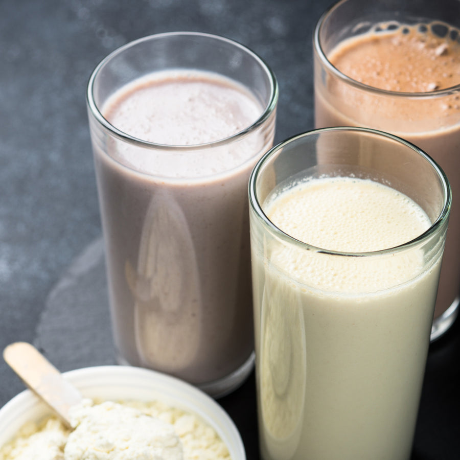 Three tall glasses of different protein shakes—light brown, dark brown, and creamy white—are arranged next to a bowl of protein powder with a scoop, set against a dark background.