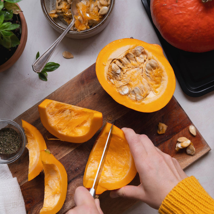 A person in a yellow sweater slices a halved pumpkin on a wooden cutting board, with pumpkin seeds and herbs nearby, and a whole pumpkin and potted plant in the background.
