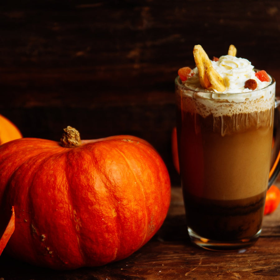 A large, bright orange pumpkin sits beside a glass of pumpkin spice latte topped with whipped cream and garnished with nuts and dried fruit, set against a rustic wooden background.