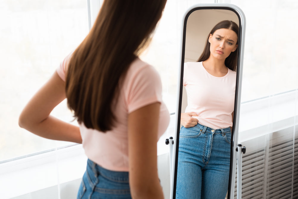 A woman in a pink shirt stands in front of a mirror, looking concerned as she examines her reflection, which shows her wearing fitted jeans in a bright, softly lit room.