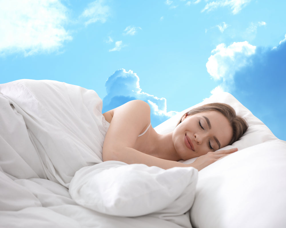 A woman with a peaceful expression sleeps on a bed surrounded by fluffy white pillows, with a bright blue sky and clouds visible in the background, symbolizing restful sleep.