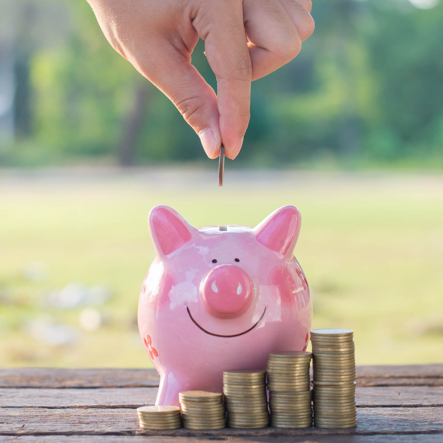 A hand is dropping a coin into a smiling pink piggy bank, with stacks of coins beside it, set against a blurred green outdoor background.
