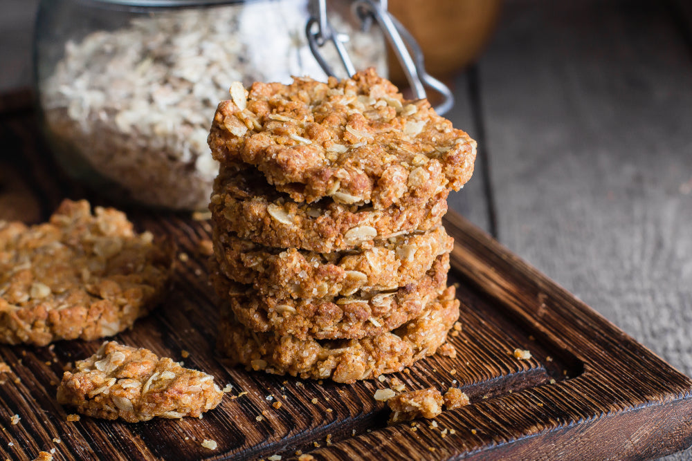 A stack of golden-brown gingerbread oatmeal cookies sits on a wooden board, with a few cookies scattered nearby and a jar of oats in the background.