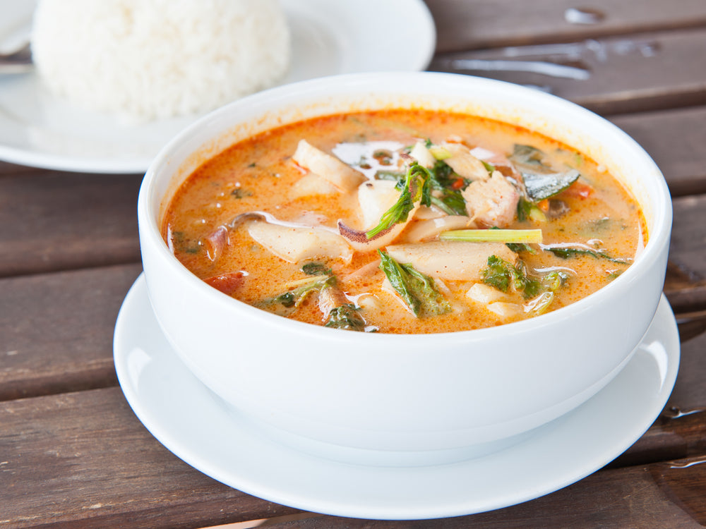 A white bowl filled with vibrant orange soup containing sliced vegetables and herbs sits on a wooden table, accompanied by a mound of white rice on a plate in the background.