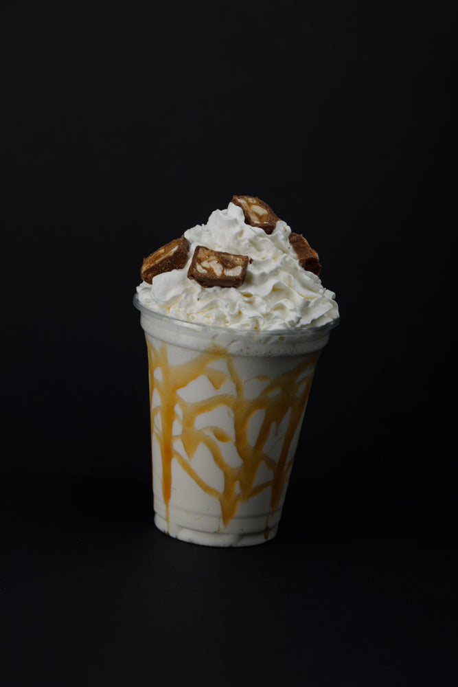 A clear cup filled with a creamy drink is topped with whipped cream and chocolate pieces, drizzled with caramel sauce, set against a dark background.