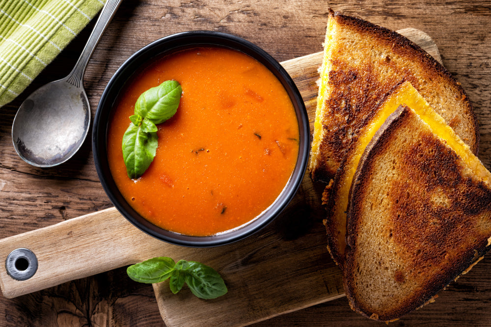A black bowl of hearty tomato soup garnished with fresh basil sits on a wooden cutting board next to three slices of golden-brown grilled cheese sandwiches and a silver spoon.